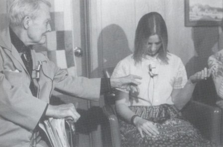 Milton Erickon con unos 70 años en su silla de ruedas, en una demostración en público. Está practicando una hipnosis con fenómeno hipnótico a Lee, una estudiante rubia de media melena,Lee es una alumna de cursos estudiante de hipnosis.