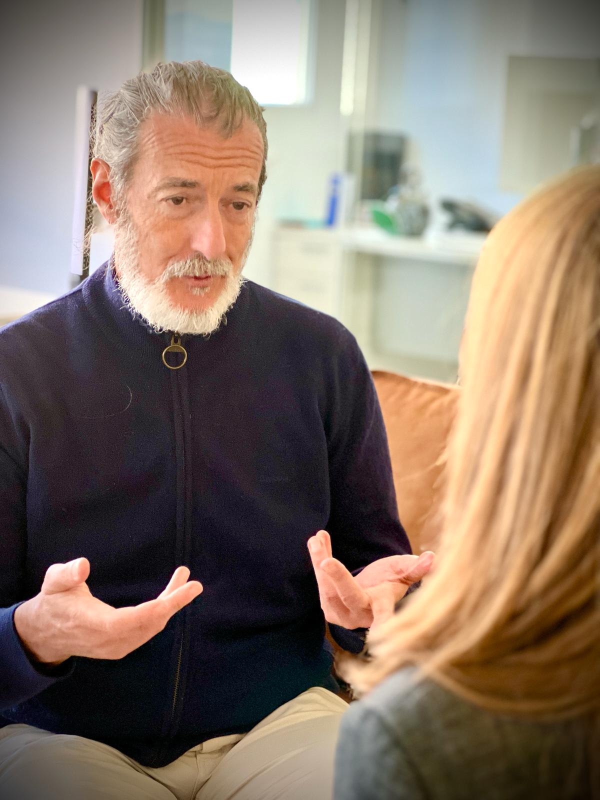 Eduardo Manchón en Hipnosis Madrid sentado en un sillón mientras mueve las manos al hablar, explicando a una paciente que está frente a él, cómo es una sesión de hipnosis.